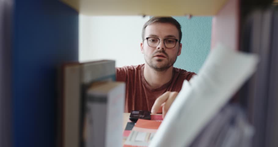 A young man browses through documents and binders on a shelf, clearly searching for something important. His expression shifts to disappointment as he fails to find what he was looking for.