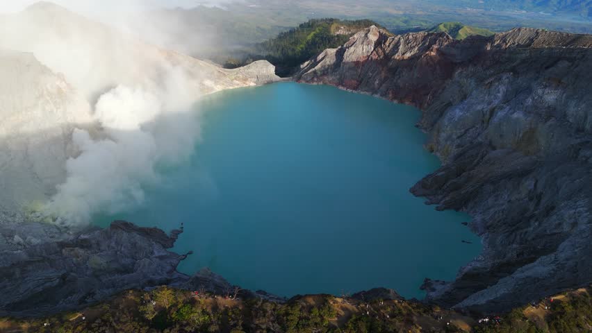 Right-orbit drone shot circles Kawah Ijen, revealing its vivid blue-green acidic lake, smoking sulfur vents and rugged caldera rim high in the mountains of East Java, Indonesia.