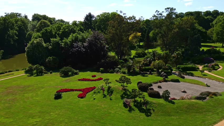 Vibrant red flowerbed design on green lawn, with beautiful Japanese garden in background, Parc de Haute Bretagne, France. Aerial drone low-altitude