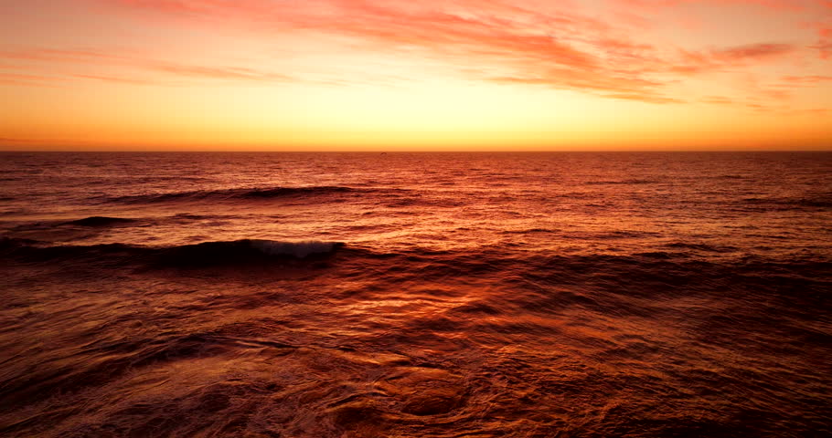 Glowing ocean waves roll under a vivid sunset sky, creating a dramatic and warm seascape with reflections of orange, red, and gold tones across the water. Slow motion aerial view