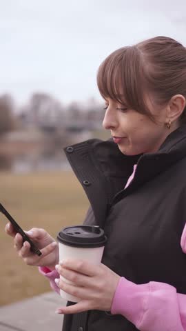 Young woman enjoys a coffee while checking her phone during a leisurely stroll through a peaceful park, embracing the tranquility of nature and the convenience of modern technology vertical video