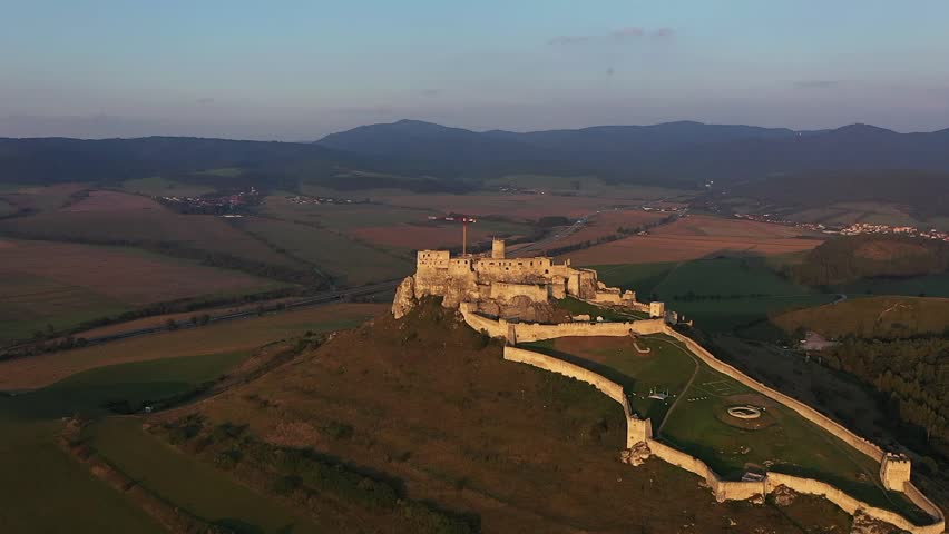A drone flies toward the illuminated Spiš Castle during sunset, revealing its stone towers, golden glow, surrounding fields, and blue-grey storm clouds.