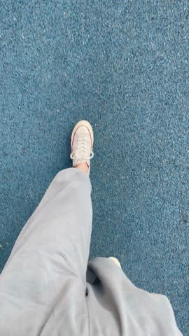 POV. First person view of a person walking on blue textured outdoor pavement, in light-colored sneakers and casual pants. Dynamic and minimal composition with a modern urban feel.