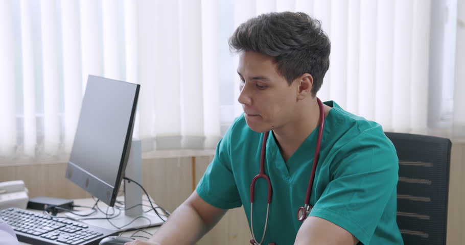 Asian male nurse concentrating during hospital consultation while listening to senior doctor about patient treatment plan using computer and medical documents in clinical office