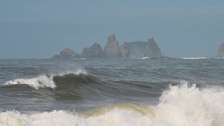 A drone captures waves rolling onto the sandy shoreline of Olympic National Park, Washington. Rocky sea stacks rise from the ocean in the distance under a clear blue sky.