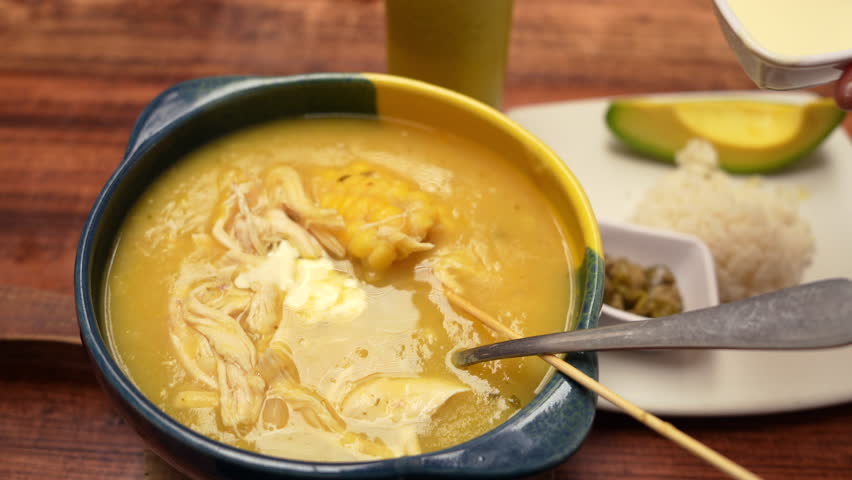 Detailed close-up shot of traditional Colombian ajiaco soup with creamy topping. Warm lighting enhances rich textures. Hand pouring cream adds a homely touch to this culinary scene.