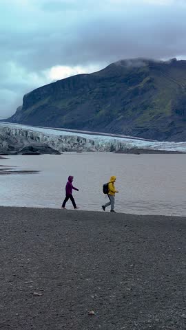Adventurers stroll along the rocky bank, taking in the majestic Skaftafell Glacier under a dramatic sky. The serene environment reflects natures beauty and power in Iceland.