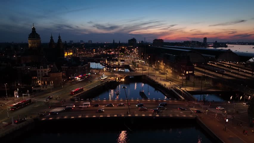 Static aerial view of traffic and reflections on Amsterdam canal bridges at night, with iconic architecture and busy roads.
