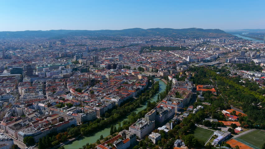 Sweeping drone footage capturing the historic cityscape of Vienna, highlighting the Danube Canal, vibrant rooftops, lush parks, and iconic landmarks under clear skies.