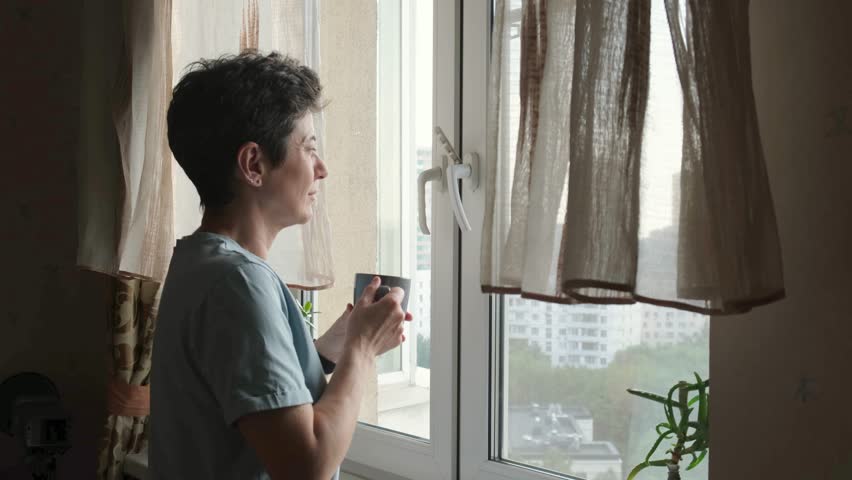 Side view of a middle-aged woman with short hair holding a cup of coffee and looking out the window, capturing a moment of relaxation and daily routine at home