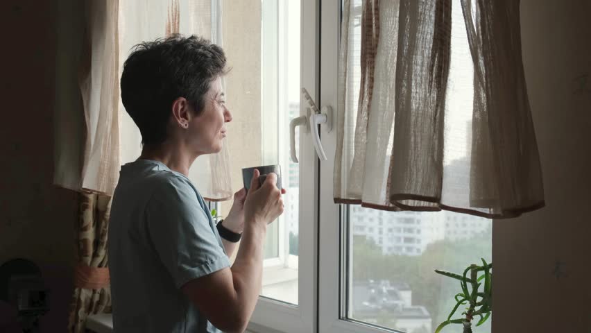 Side view of a middle-aged woman with short hair holding a cup of coffee and looking out the window, capturing a moment of relaxation and daily routine at home