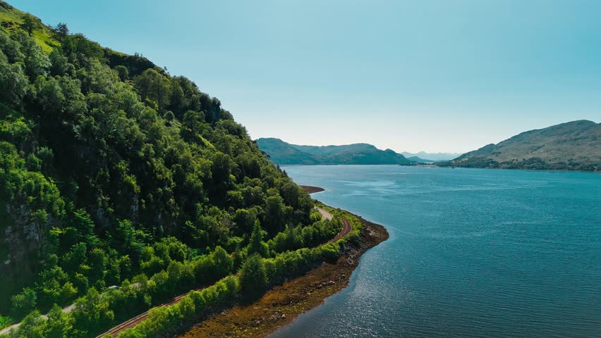 This aerial view captures the lush green hills meeting the tranquil waters of the loch in Scotland, showcasing the natural beauty of the landscape under clear skies.