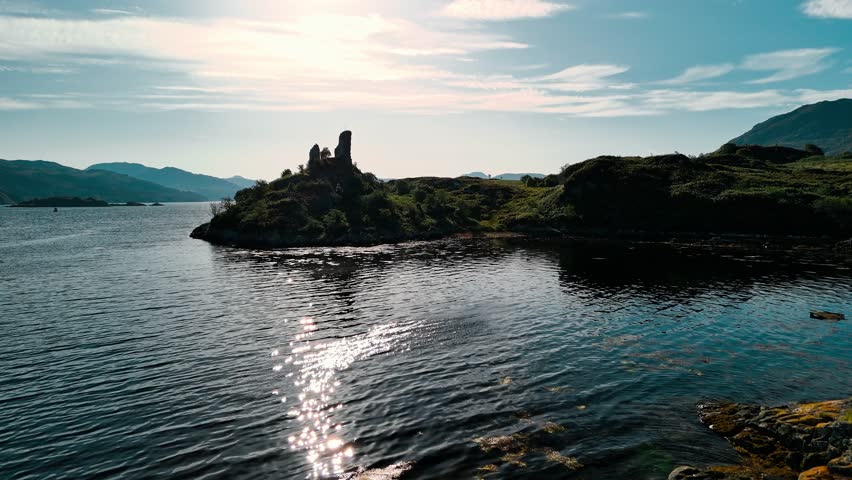 This aerial view shows a castle ruin on a small island, surrounded by calm waters and mountains, highlighting Scotland's stunning natural beauty.