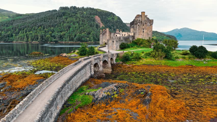 Eilean Donan Castle stands majestically against a backdrop of green hills and water. The stone bridge leads visitors to this iconic Scottish site, rich in history.