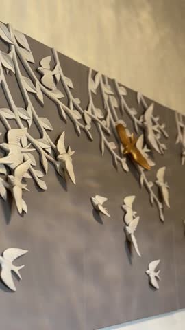 Close-up of a decorative wall panel with sculpted birds and branches, some highlighted in gold, against a light gray backdrop.