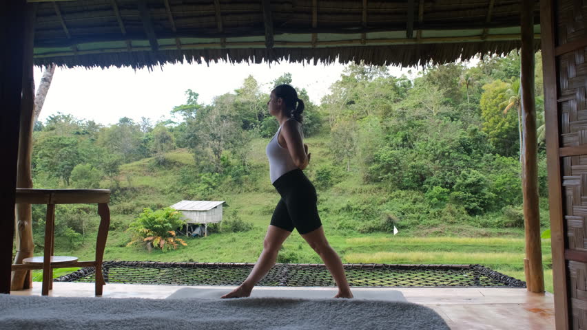A young woman engages in a yoga pose on a terrace of bamboo hut, surrounded by vibrant tropical foliage and lush rice plains, slow motion. Active and healthy lifestyle
