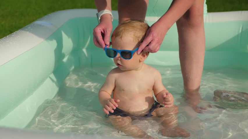 A cute baby boy happily plays in a small inflatable pool. His mother carefully puts on stylish sunglasses for him