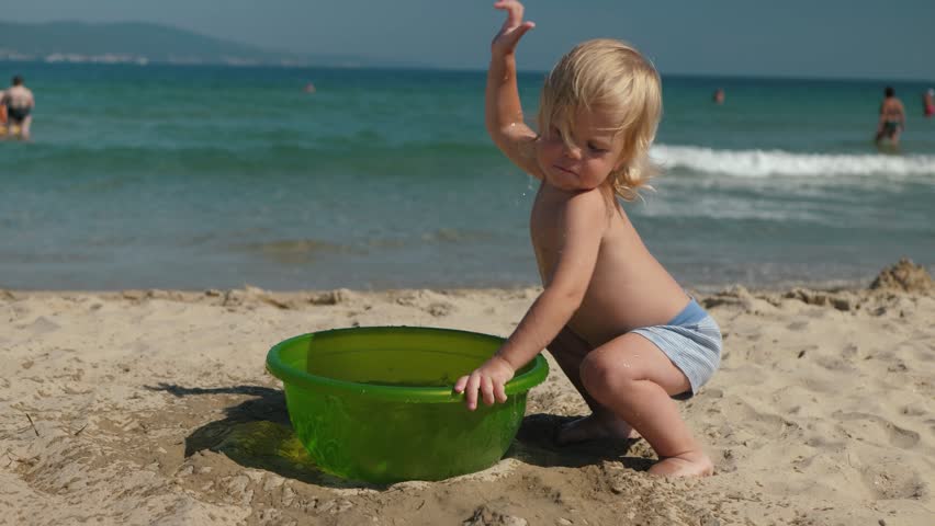Joyful little toddler happily plays by the seaside, splashing water enthusiastically from a bright green bath basin onto the warm sandy beach, capturing the true essence of summer fun and joy