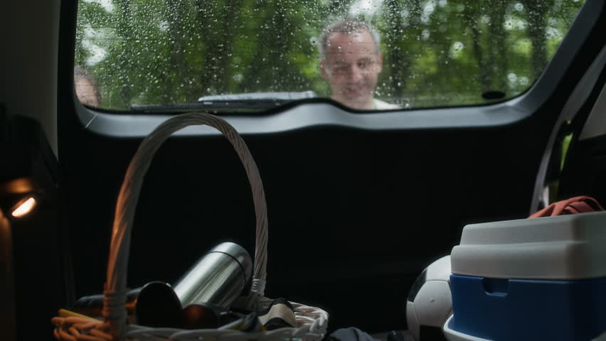 Family of travelers opening car trunk and taking out backpacks, basket and box with picnic items, soccer ball and two badminton rackets, then shutting back door of vehicle