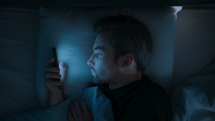 Close up, a tired man in bed scrolls endlessly on his mobile phone, struggling with insomnia, anxiety and overthinking as he watches social media late into the night. Smartphone dependence. Top view