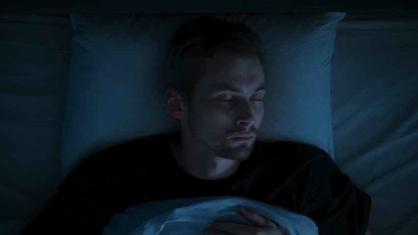 Close-up, in a dark bedroom at night, a man wakes up because of sleep paralysis or nightmare, drained by insomnia and a sleep disorder, overwhelmed by anxiety, fatigue.