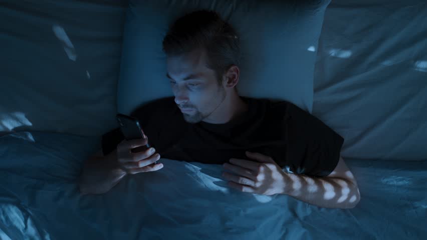 Top view, young man laying in bed at night, holding smartphone and doom scrolling through social media, then tossing the phone aside in exhaustion and frustration. Smartphone addiction and insomnia