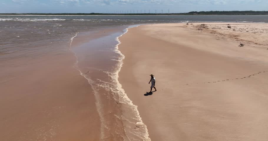 Man in white clothes walks on a curved sandbar beside muddy Parnaíba River waters in Araioses, Brazil. Aerial follow shot.
