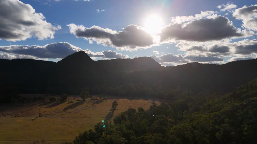 Aerial view of sun setting behind mountain ridge, casting dramatic light over rolling hills and forest in rural New South Wales, Australia