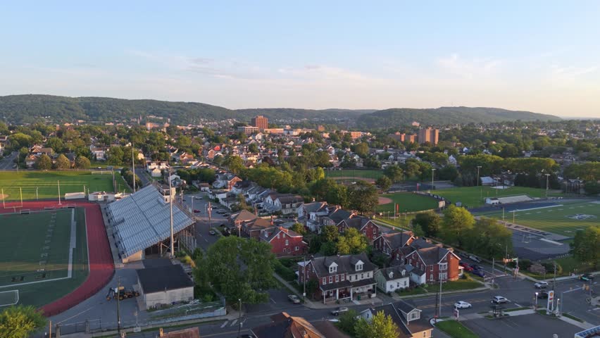 Aerial flyover of Bethlehem, Pennsylvania with sports stadiums, houses, and distant hills under warm sunset light. Dolly in