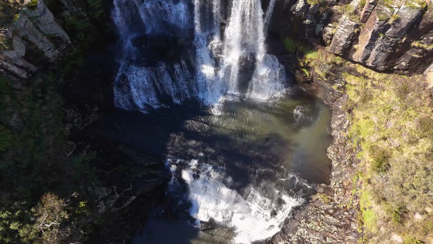 Drone footage captures cascading waterfall and faint rainbow over rocky gorge at Ebor Falls, New South Wales, under bright natural daylight with minimal camera movement