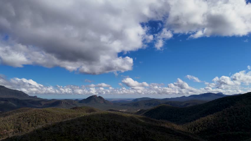 Drone camera smoothly pans across rugged Warrumbungle mountain landscape under dramatic clouds and late afternoon sunlight, revealing expansive Australian wilderness in vivid detail