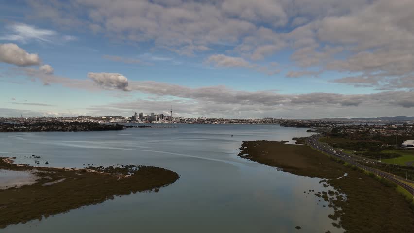 A scenic 4K 60fps aerial flight over the tranquil waters of Shoal Bay, revealing the stunning Auckland CBD skyline, including the Sky Tower, in the background. A perfect shot of NZ city life