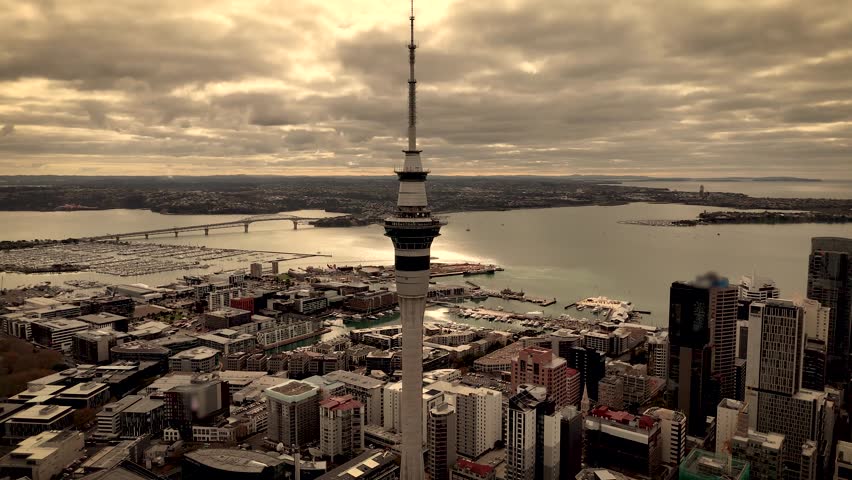 Iconic Sky Tower at golden hour, CBD, Harbour Bridge, and Waitematā Harbour under cloudy sky, North Island, New Zealand. Aerial drone orbiting