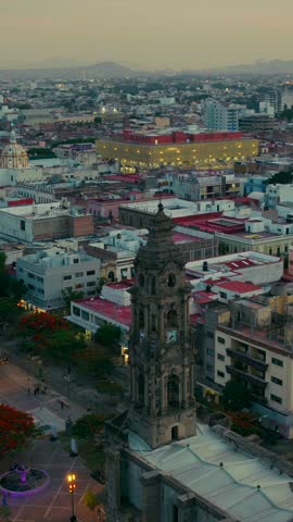 Templo San José de Gracia bell tower. Reveal orbit shot of carousel, illuminated spires of the Guadalajara Cathedral, Avenida Fray Antonio Alcalde and historical city center. Vertical shot