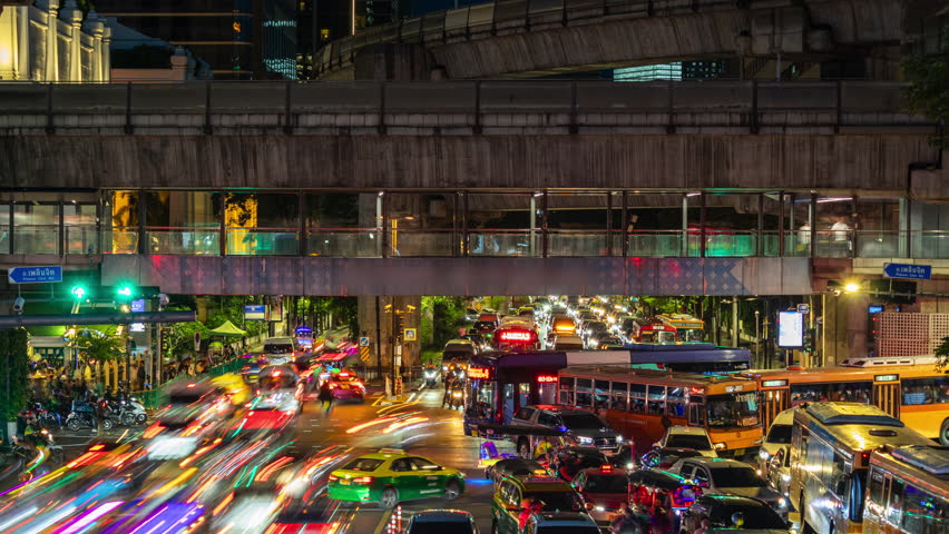 Vibrant nighttime traffic scene in Bangkok. Cars and motorcycles navigate busy intersections beneath elevated train tracks. Colorful lights reflect on the asphalt, revealing city life
