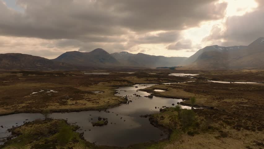 Aerial drone footage of the dramatic Glencoe valley in the Scottish Highlands, captured in moody pre-evening light. Sweeping views of rugged mountains, winding roads, and wild, untouched nature