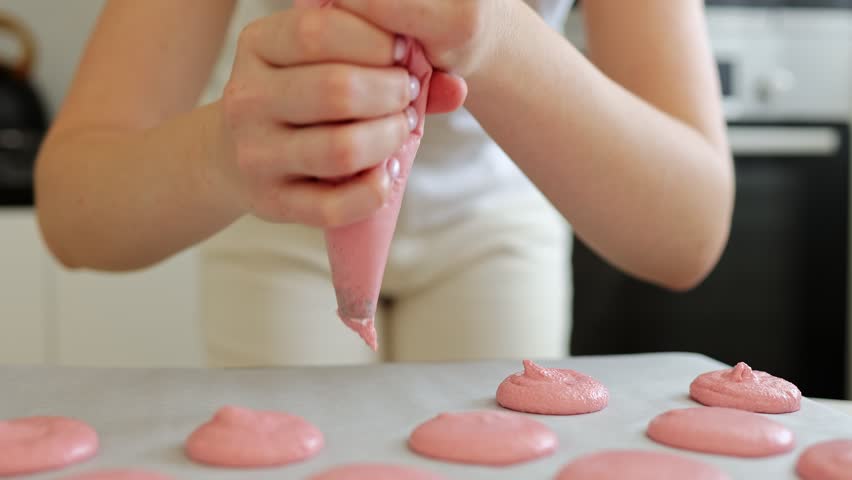 hands expertly pipe vibrant pink macaron batter onto parchment paper