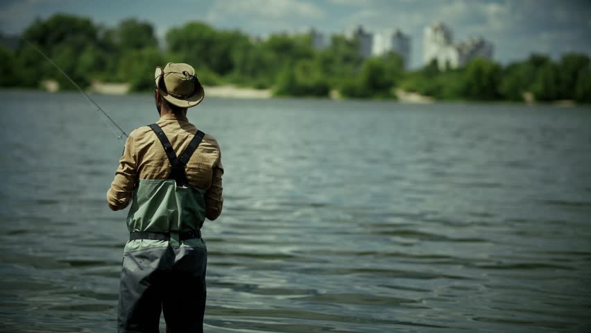 Man Fly Fishing In Calm Lake Waters.  Back View Of Angler With Rod. Outdoor Recreation Activity Peaceful Nature Retreat. Summer Fishing Experience Solitary Hobby. Fisherman In Waders And Fishing Gear
