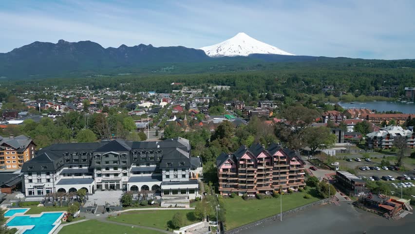 Panoramic drone view of downtown Pucón, with the snow-covered Villarica volcano in the background and scenic architecture along the lake shore.