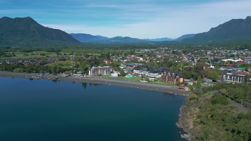 Orbiting drone view of Pucón’s beach and town, with calm lake waters, hotels, and scenic mountains surrounding the area.