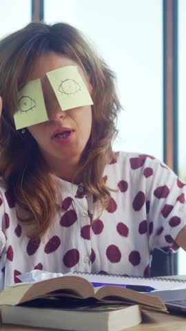 Woman adds humor to her study session by placing sticky notes on her eyes. She seems to enjoy a lighthearted moment while sitting at a table full of books.
