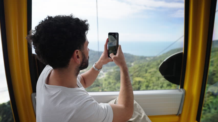 Man taking photo as tourist from inside moving cable car over forested hill environment, Balneário Camboriú, Santa Catarina, Brazil.
