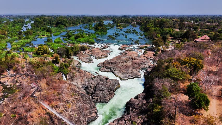 Drone pulls away over turbulent Mekong rapids near Don Det, Laos, revealing fractured basalt flows, rocky fault lines, and scattered islets across the braided floodplain in uplifted Champasak Plateau.