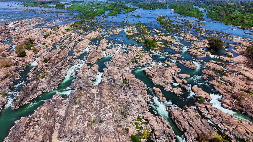 Wide top-down drone shot over Mekong River near Don Det, Laos, reveals interwoven rapids cutting through jagged rock beds and narrow channels across the fractured floodplain of the Si Phan Don region.