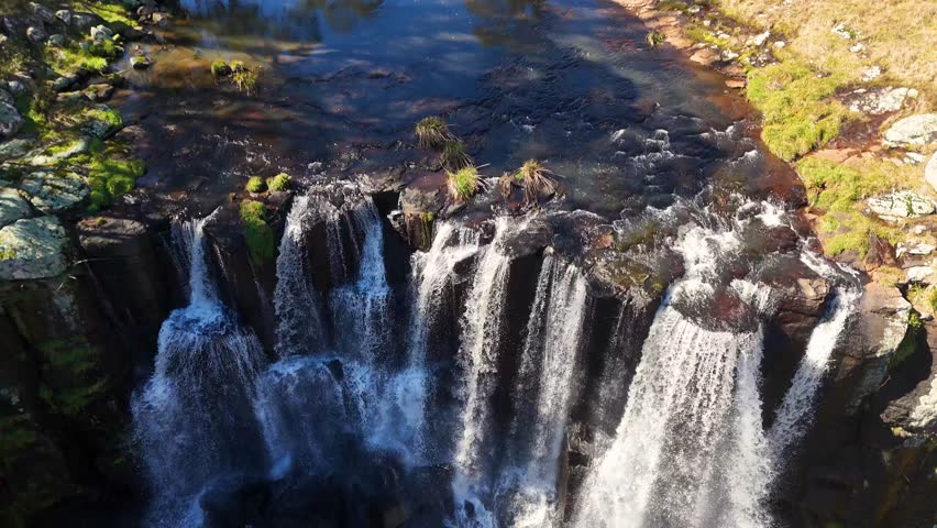 Drone footage captures cascading waterfalls at Ebor Falls, New South Wales, Australia. Bright daylight highlights lush greenery, rocky cliffs, and flowing water from a high, top-down perspective