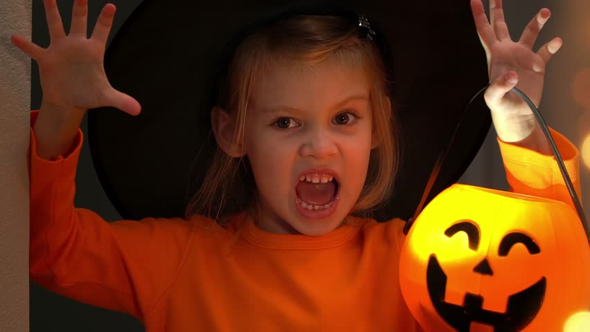 Portrait of angry little girl in witch costume and black hat fun scares with lantern pumpkin in dark. Child celebrates halloween
