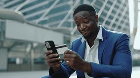 Portrait of happy African American businessman uses banking app on smartphone in urban district. Young entrepreneur pays with internet technology sitting on parapet near skyscraper - Powered by Shutterstock - Get 15% off with code: PIKWIZARD15
