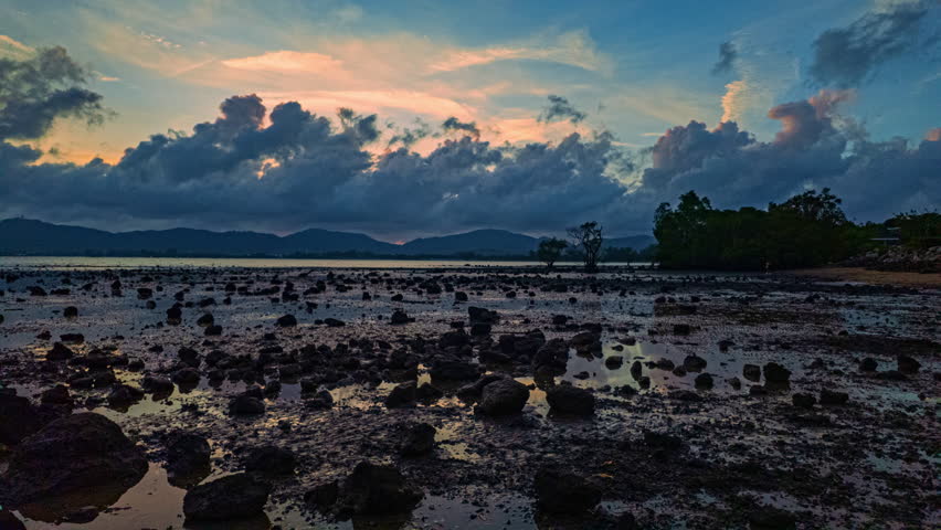 Time Lapse A dramatic twilight scene of a rocky shoreline during low tide, with silhouetted mountains and trees under a moody, cloud-filled sky. The wet ground reflects the fading light