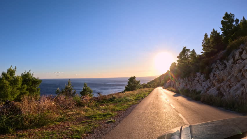 LENS FLARE: Coastal asphalt road winds along a scenic hillside, bathed in golden light of rising sun. Memorable summer morning drive through picturesque Mediterranean landscape above Adriatic Sea.