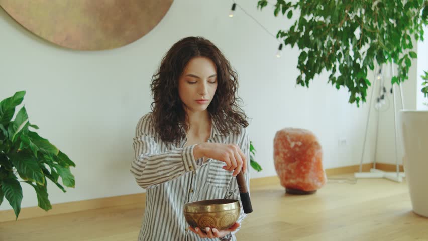 Woman using singing bowl for meditation in a bright room surrounded by plants and natural decor